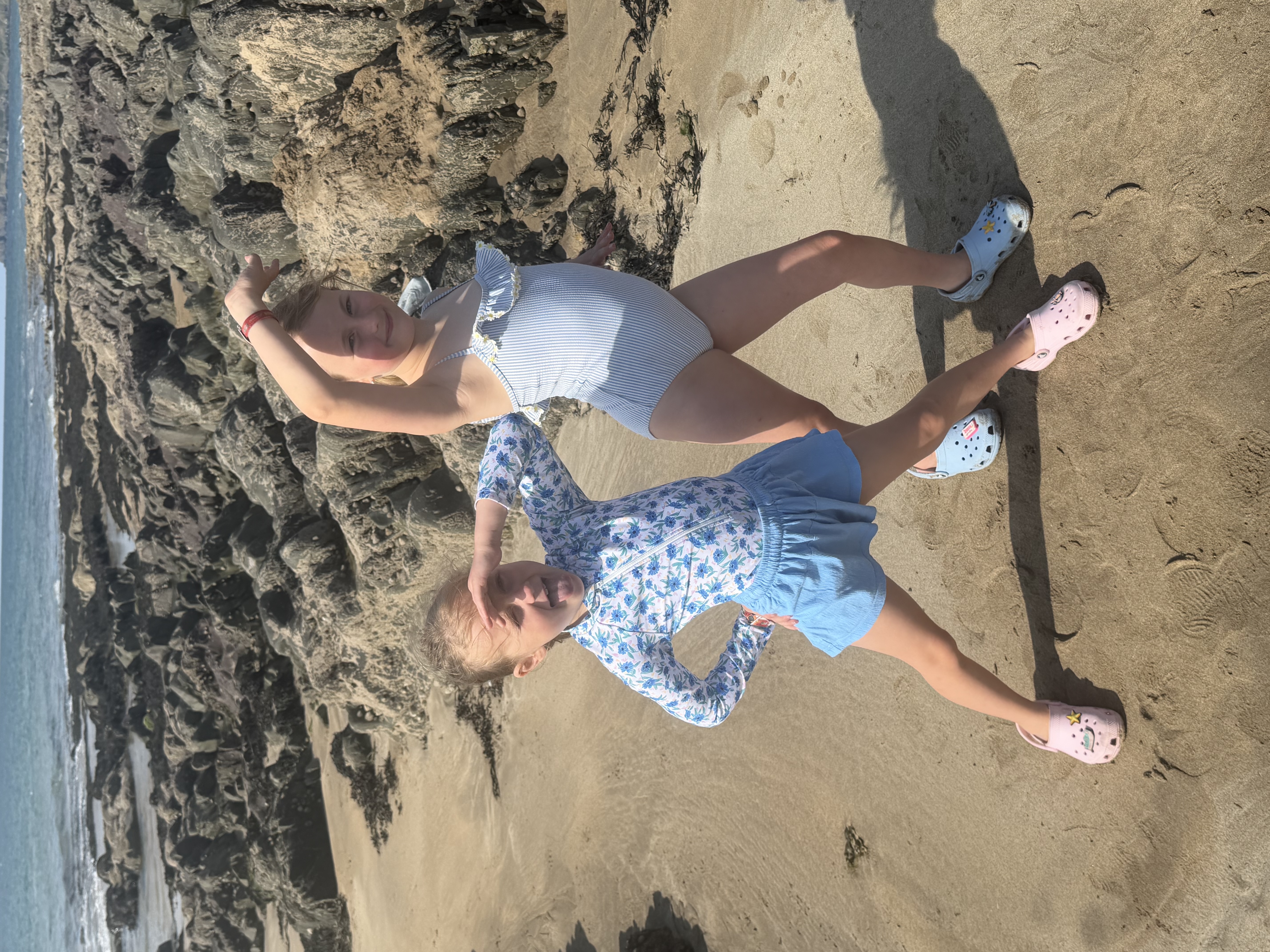 Two young girls striking playful poses on a sandy beach with rocky outcrops and the ocean in the background.