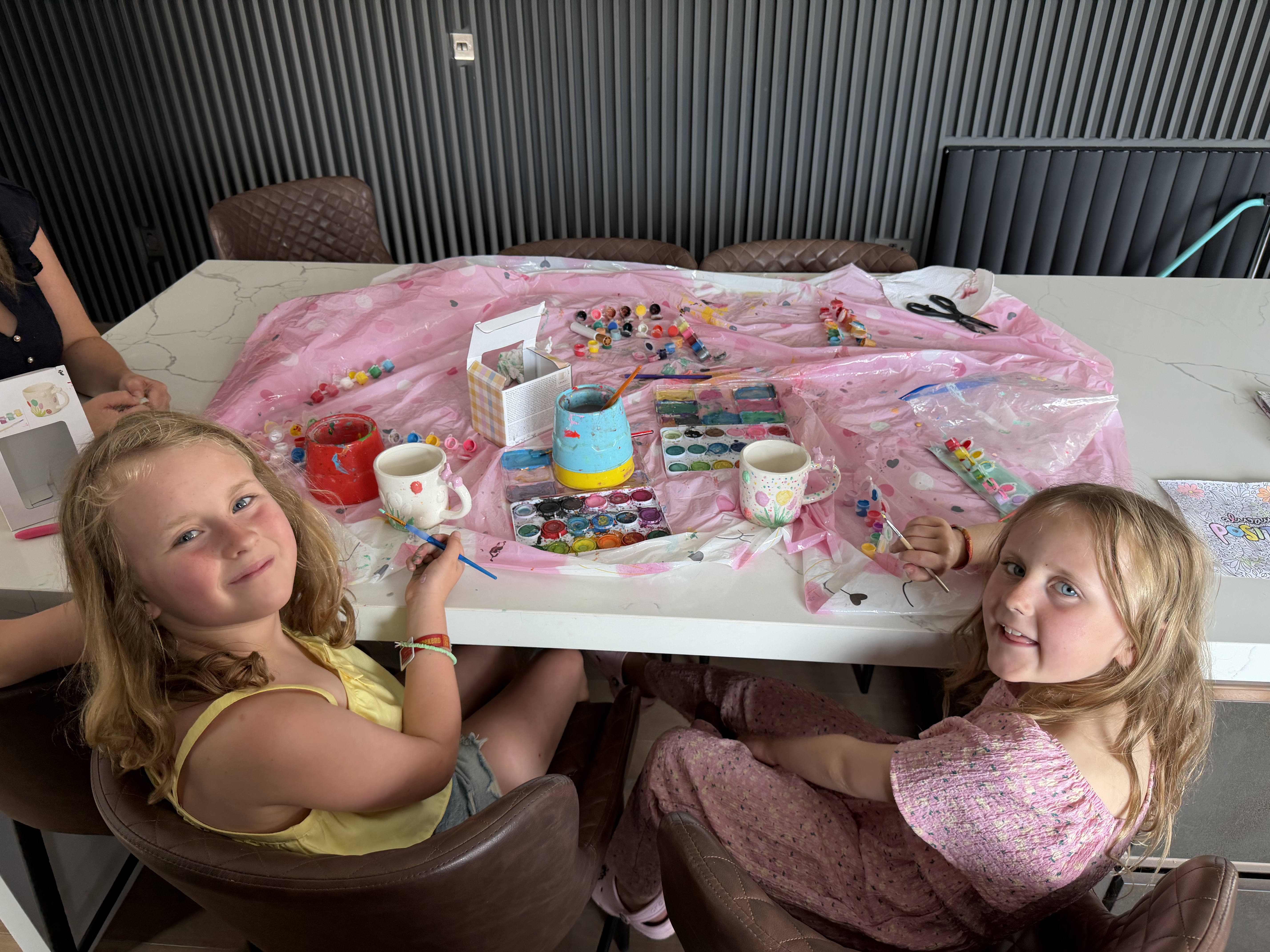 Two young girls painting ceramic mugs at a table covered with a pink plastic sheet, surrounded by paints and brushes.