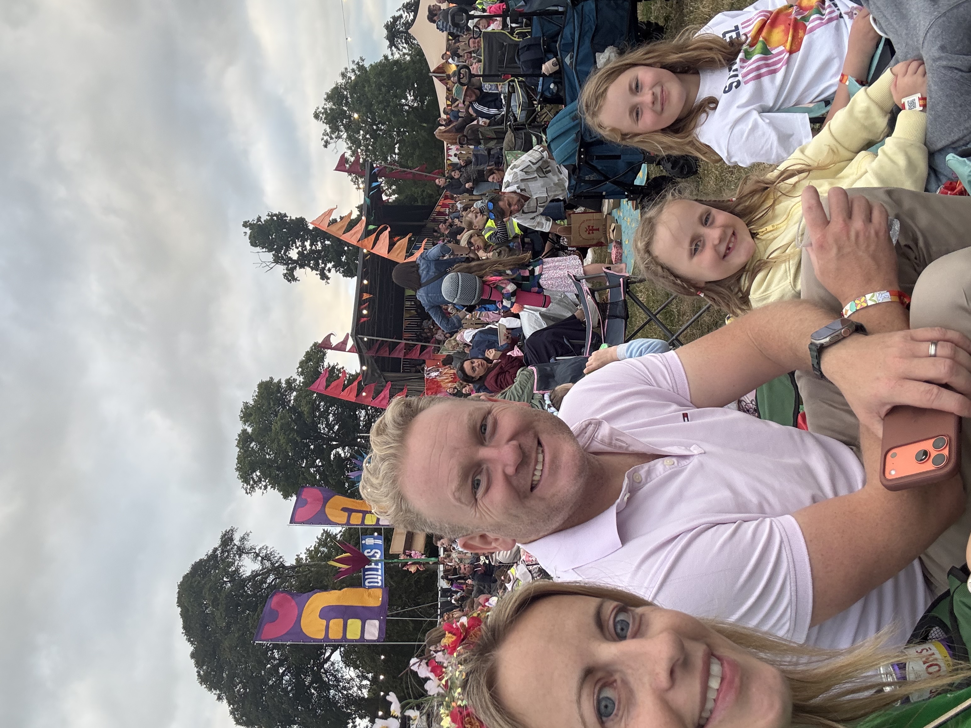 A family of four smiling at the camera at an outdoor festival with a stage and flags in the background.