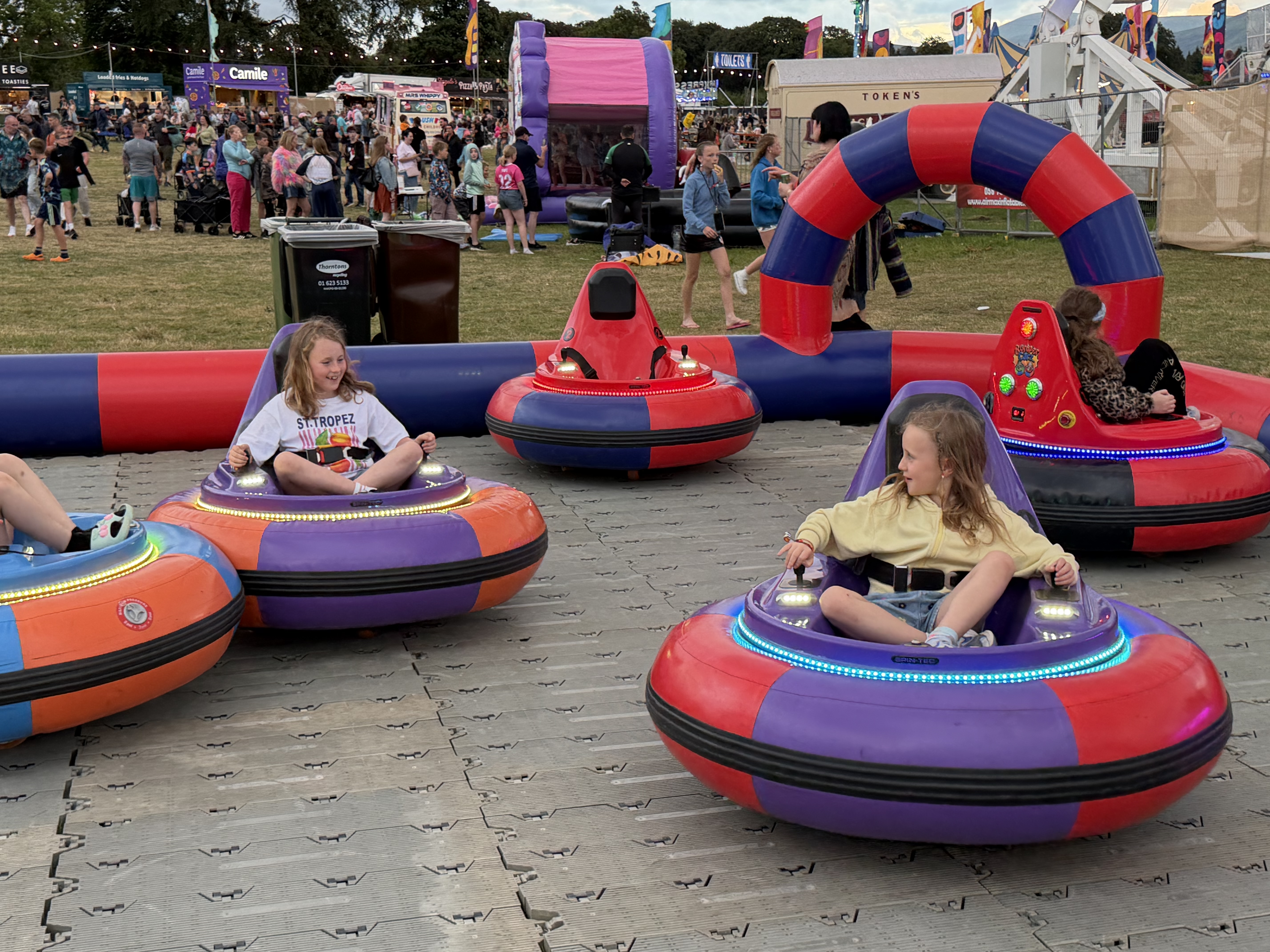 Two young girls in colorful bumper cars at an outdoor festival, with other people and tents in the background.