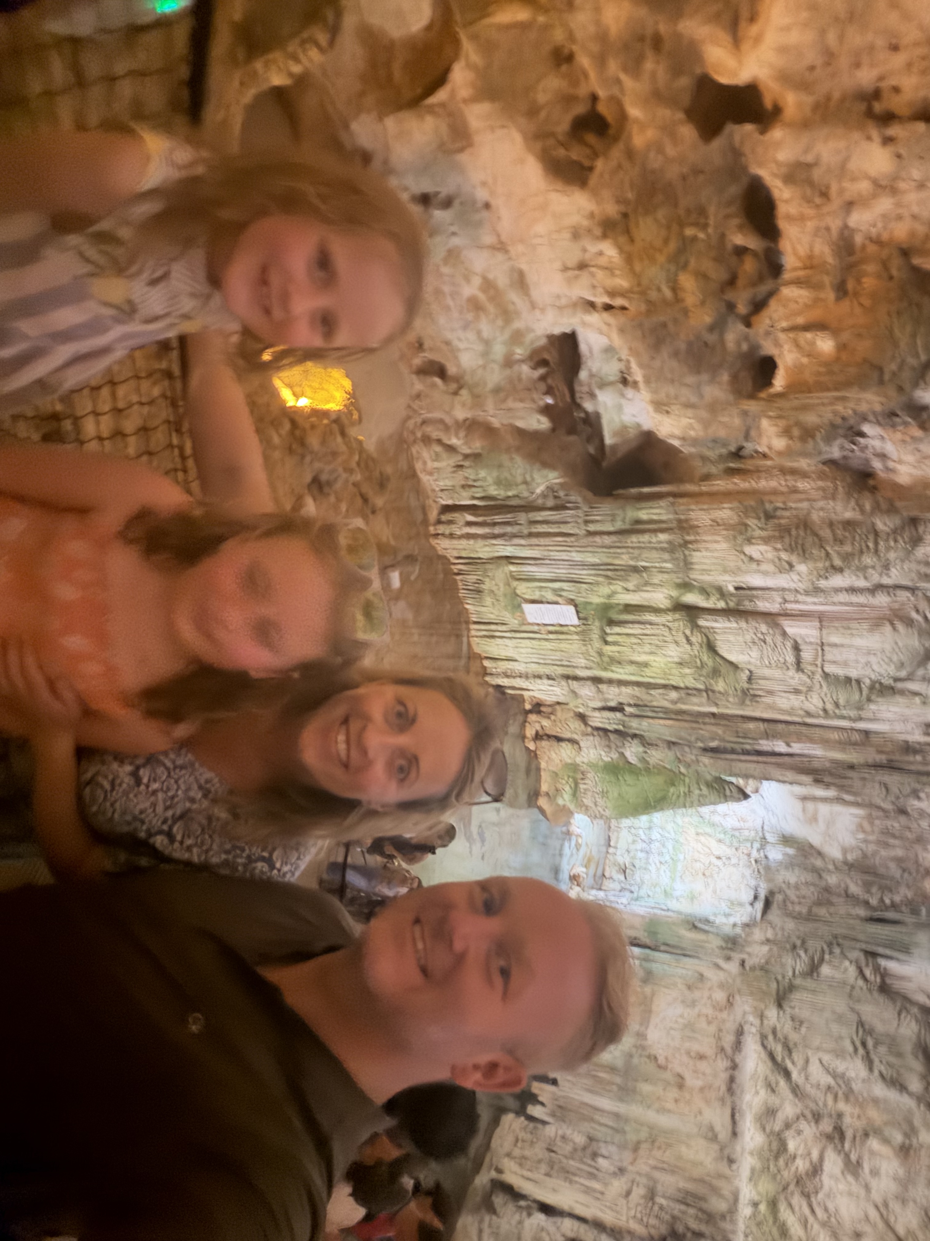 A family of four, two adults and two young girls, smiling at the camera inside a large, illuminated cave with stalactites and stalagmites.